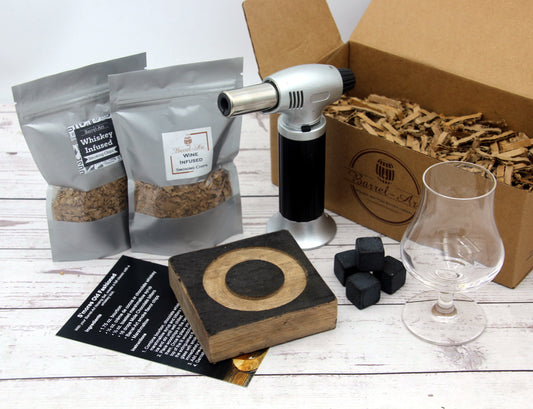 A Smoked Whiskey Cocktail Kit with a whiskey smoking tray, two bags of smoking chips, black basalt whiskey chilling stones, a butane torch, and optional glasses including a rocks glass and a nosing glass.
