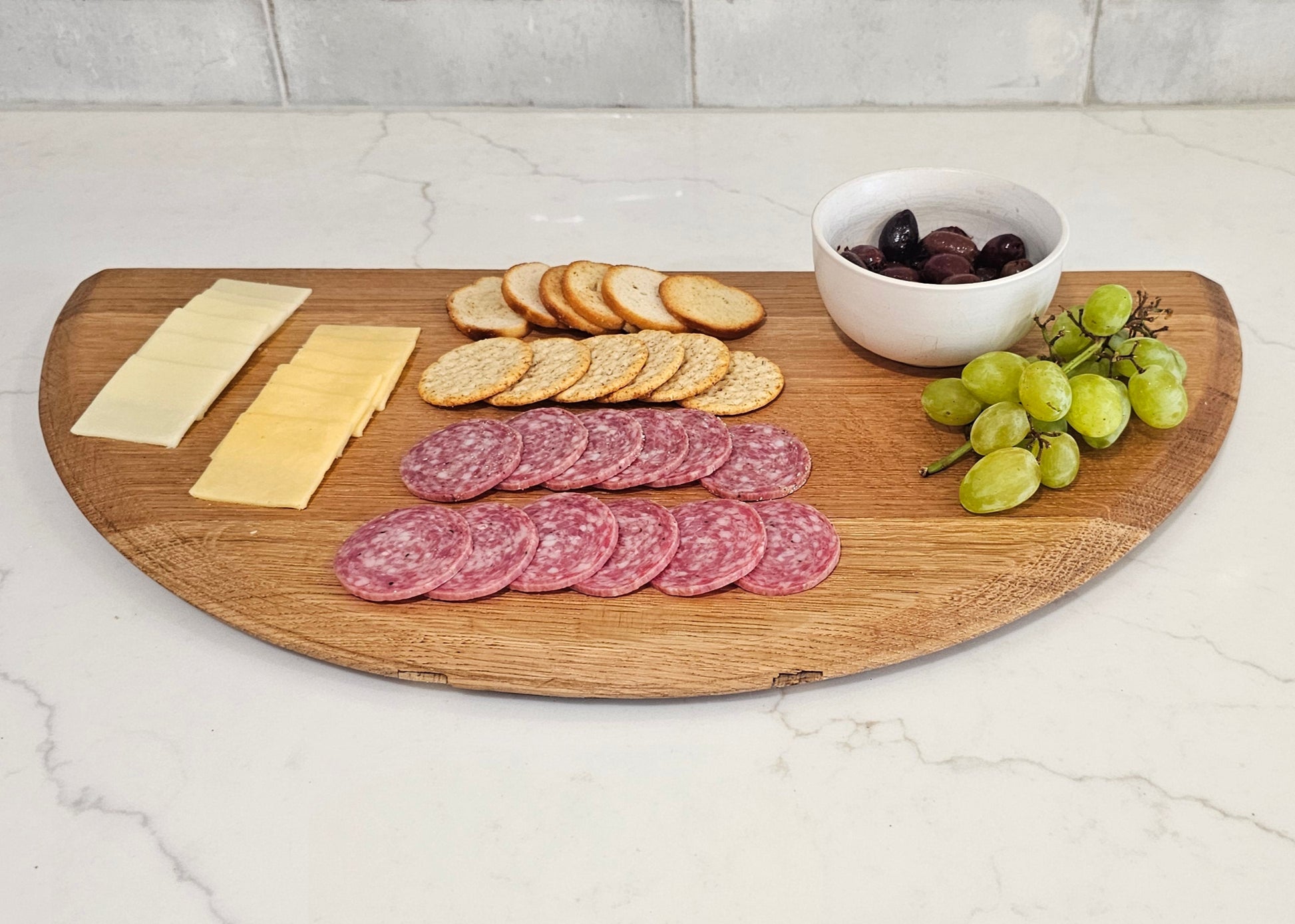 Wooden cutting board with sliced meats, cheese, crackers, and grapes on a kitchen counter.