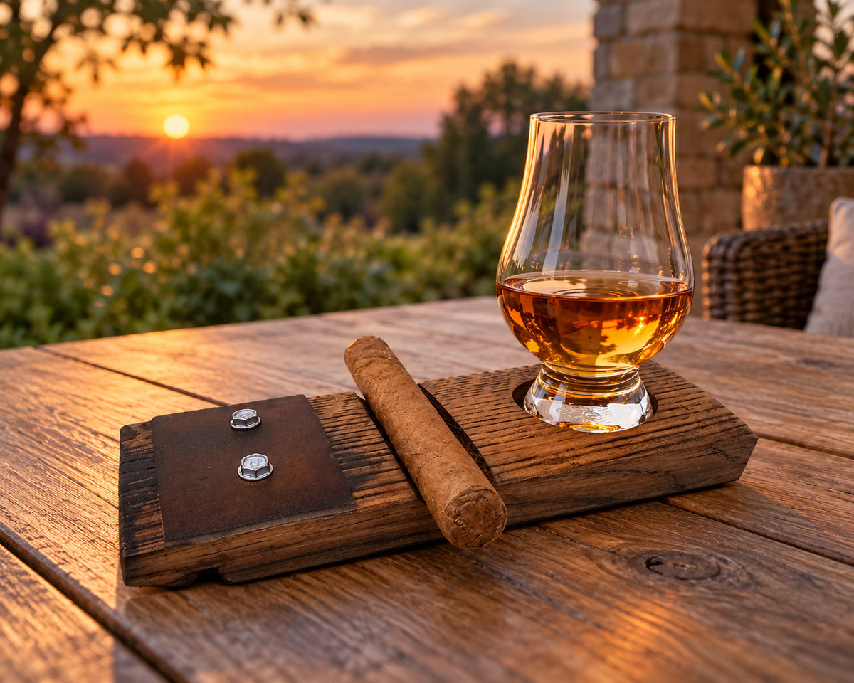 Barrel-stave glass and cigar holder on a covered patio table with a small whiskey pour and an unlit cigar.