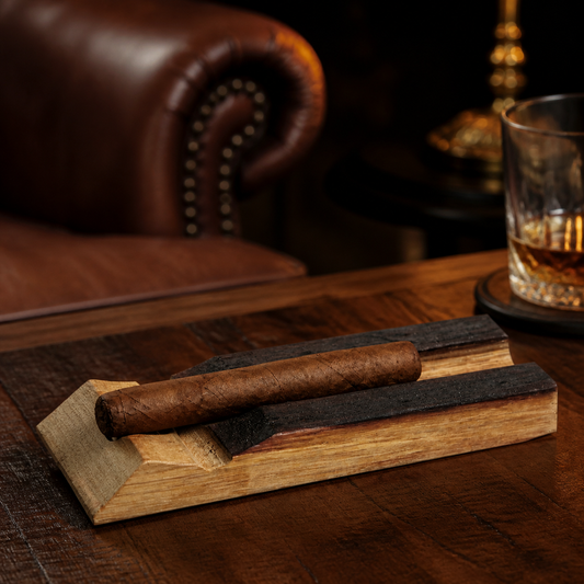 Reclaimed whiskey barrel stave cigar holder with cigar resting in charred groove on a wood table in a warm cigar lounge with whiskey glass nearby