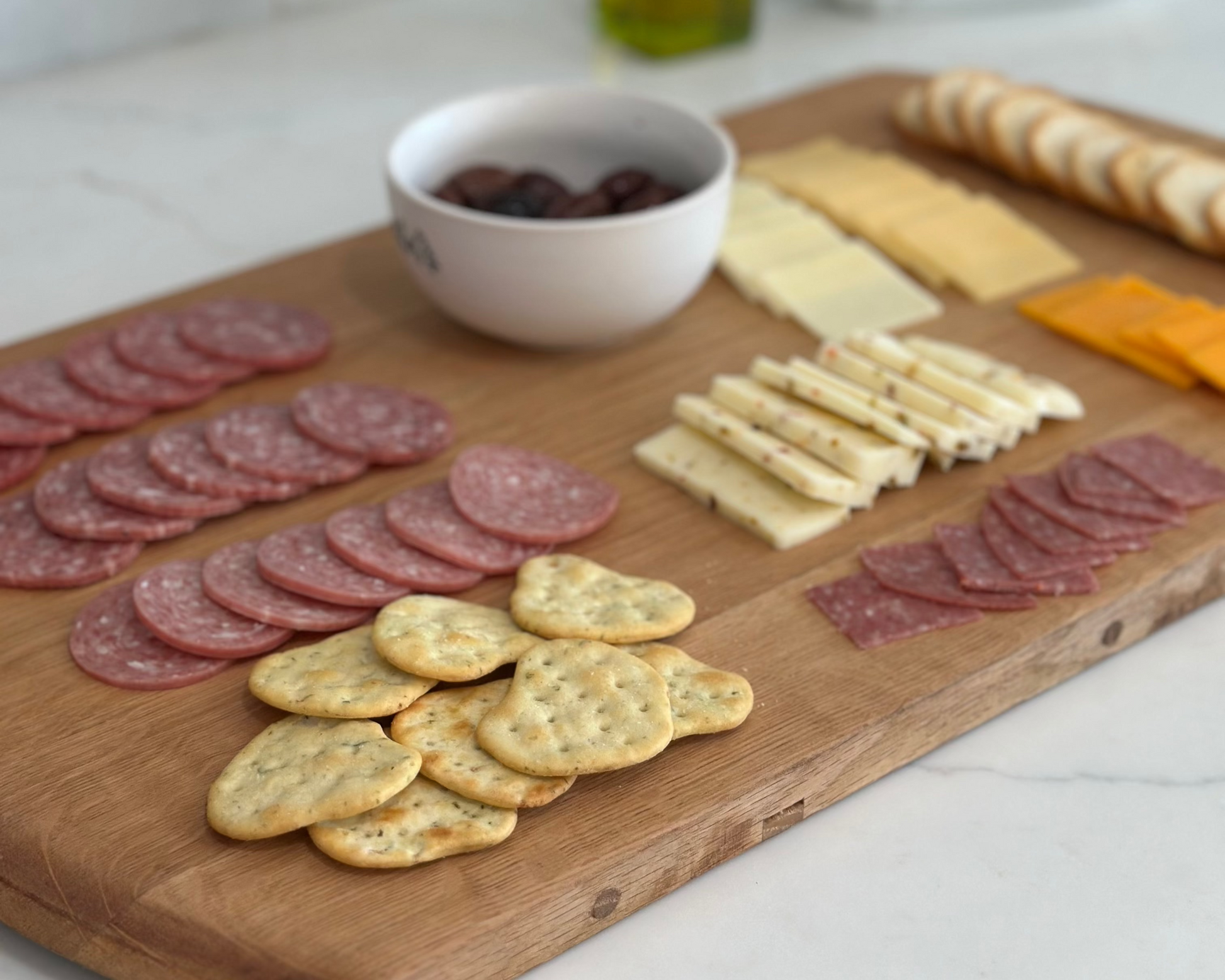 Wooden cutting board with sliced meats, cheeses, and crackers on a kitchen counter.