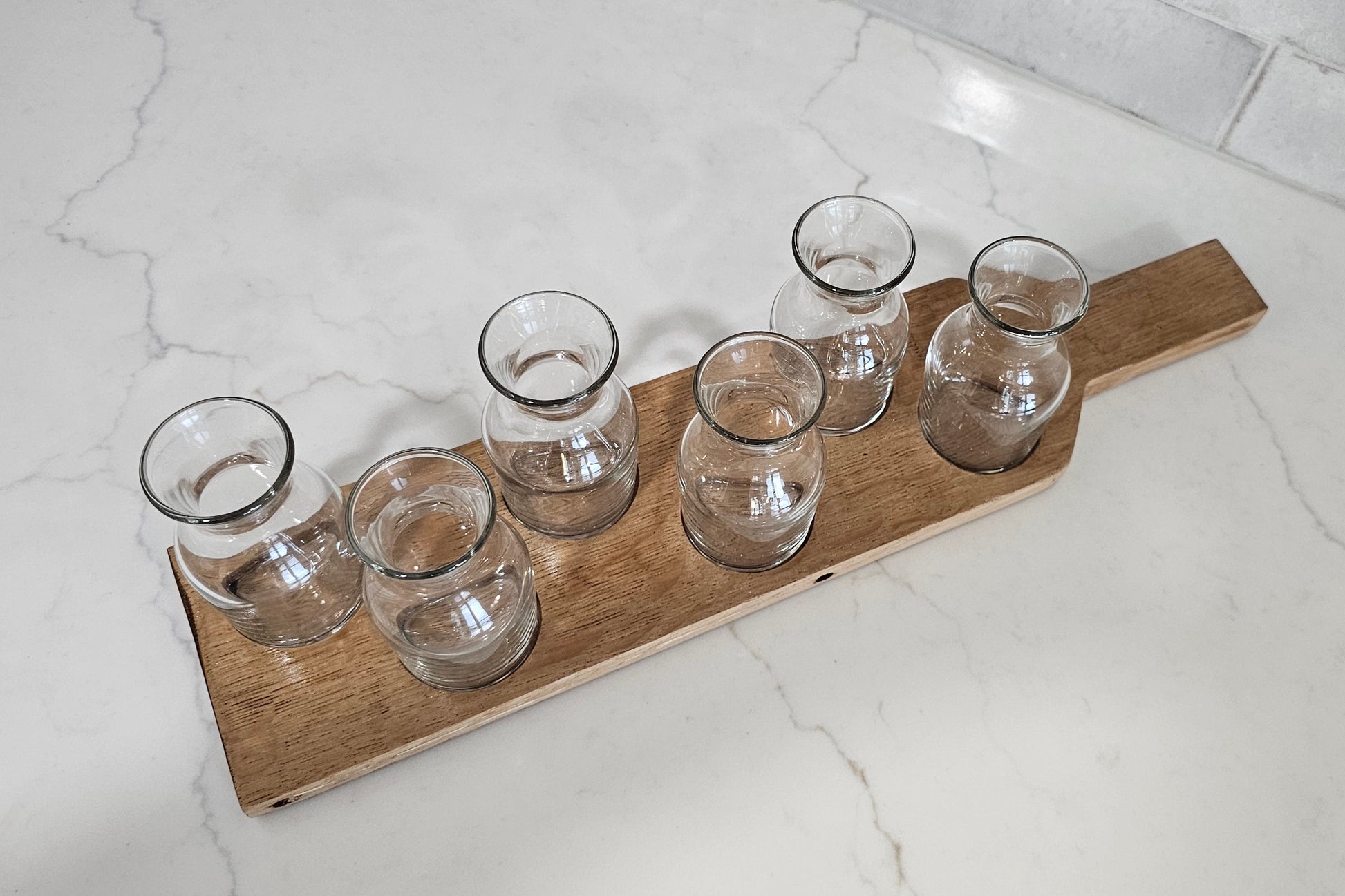 Set of six clear glass tumblers on a wooden tray against a white tiled wall.