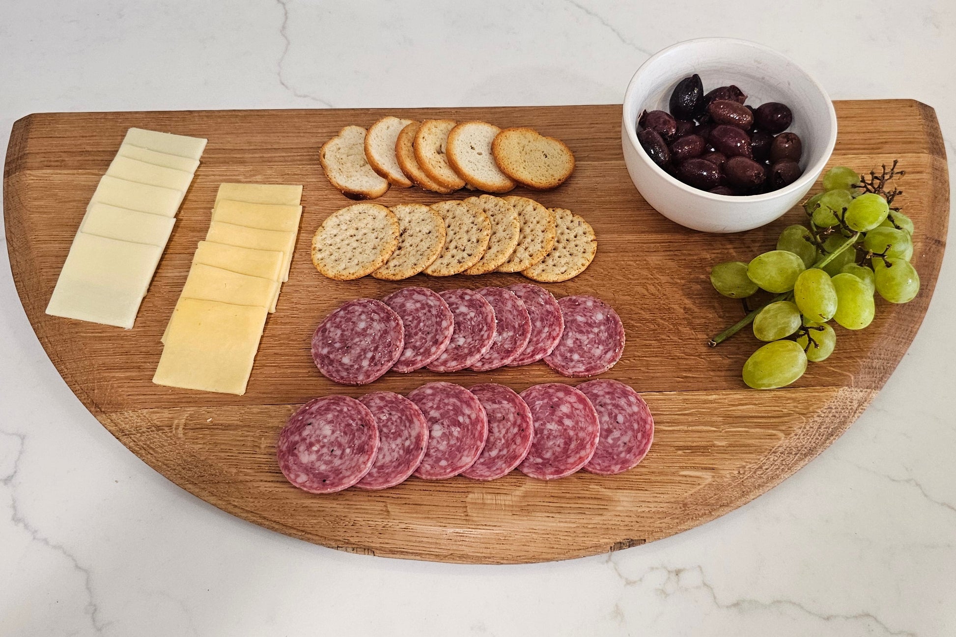 Wooden cutting board with sliced salami, cheese, crackers, and grapes on a marble surface.