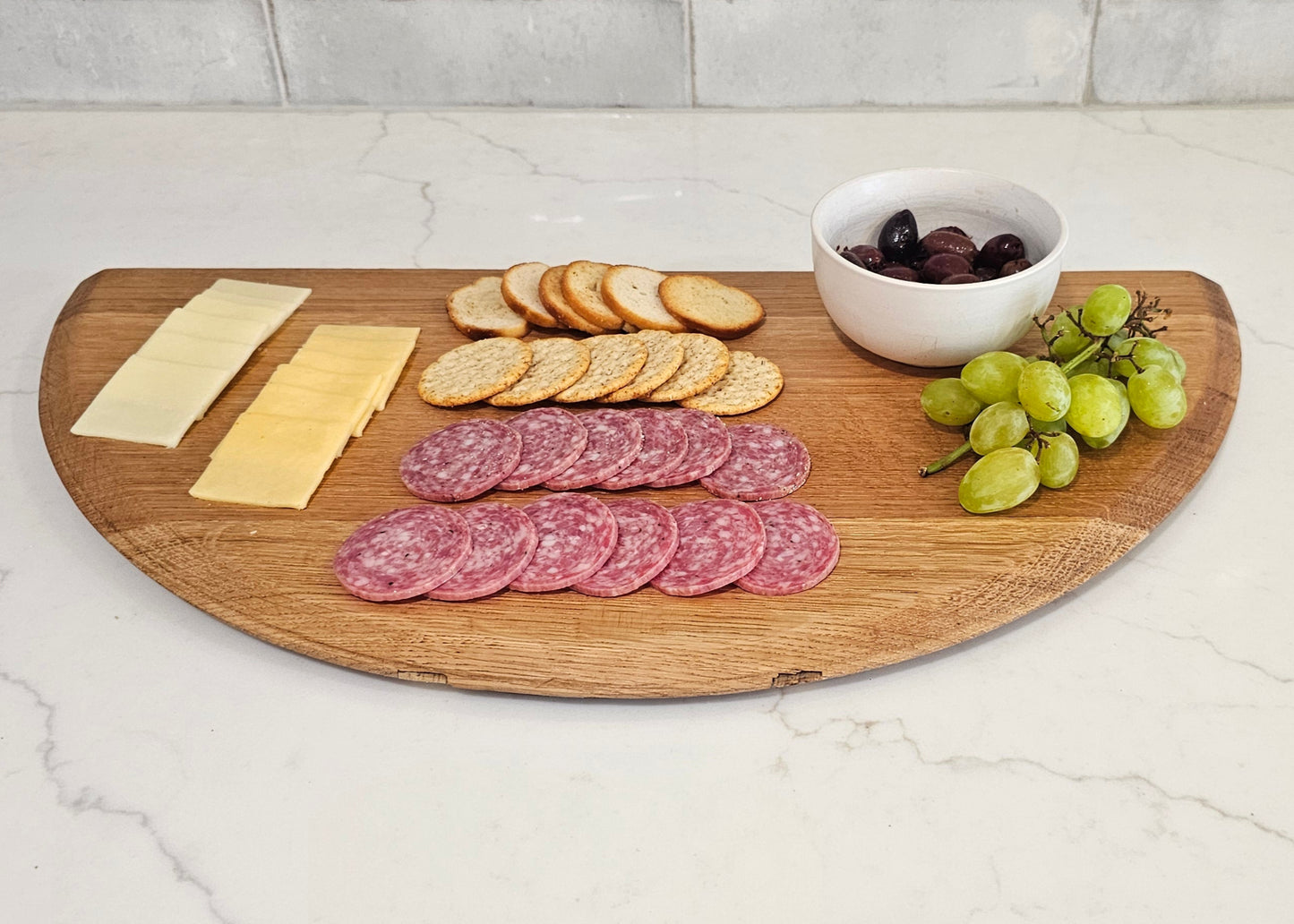 Wooden cutting board with sliced meats, cheese, crackers, and grapes on a kitchen counter.