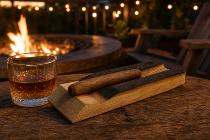 Whiskey barrel cigar holder with cigar on a rustic outdoor patio table beside a whiskey glass near a firepit and string lights