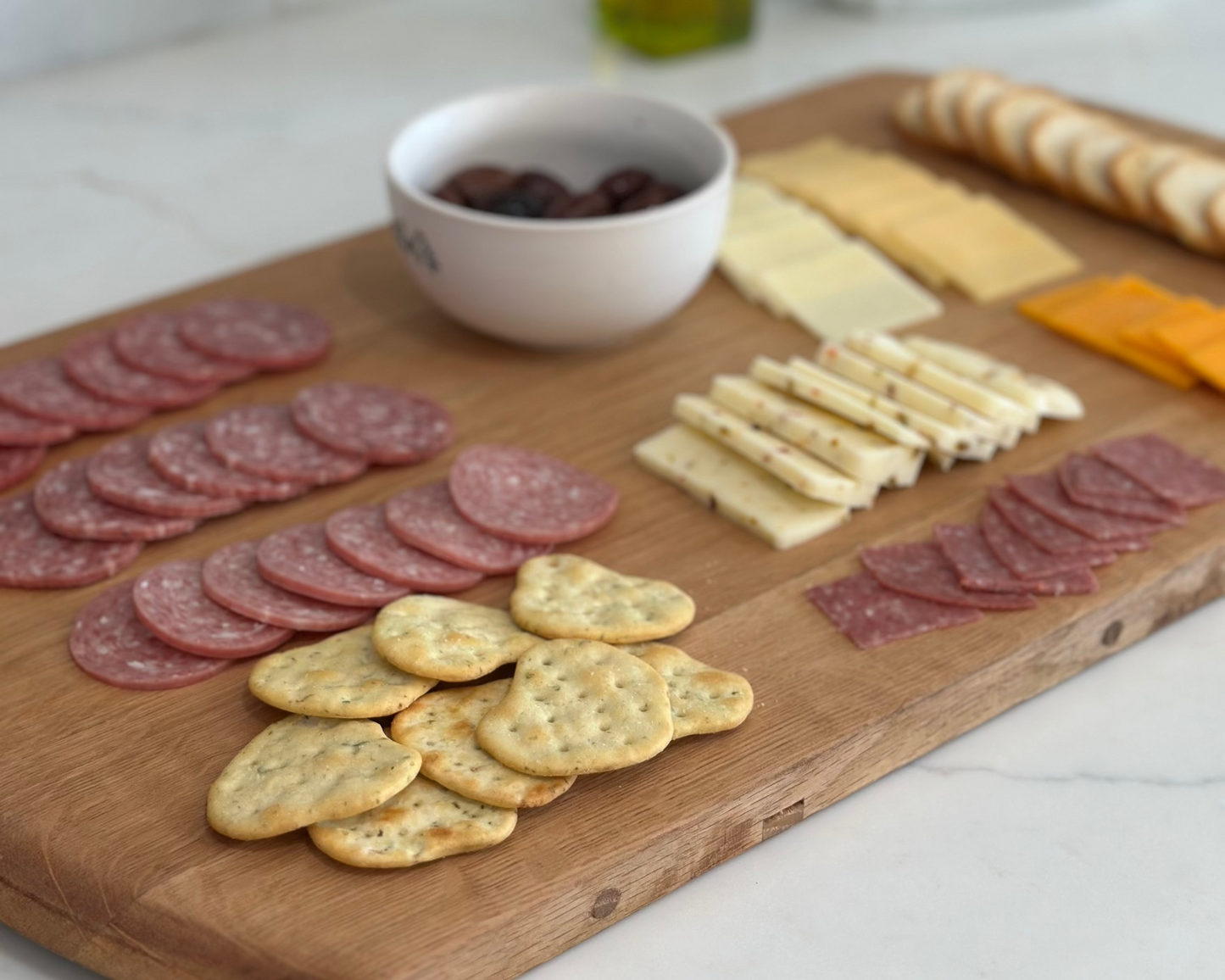 Wooden cutting board with sliced meats, cheeses, and crackers on a kitchen counter.