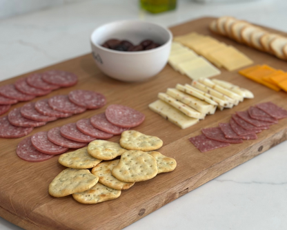 Wooden cutting board with sliced meats, cheeses, and crackers on a kitchen counter.